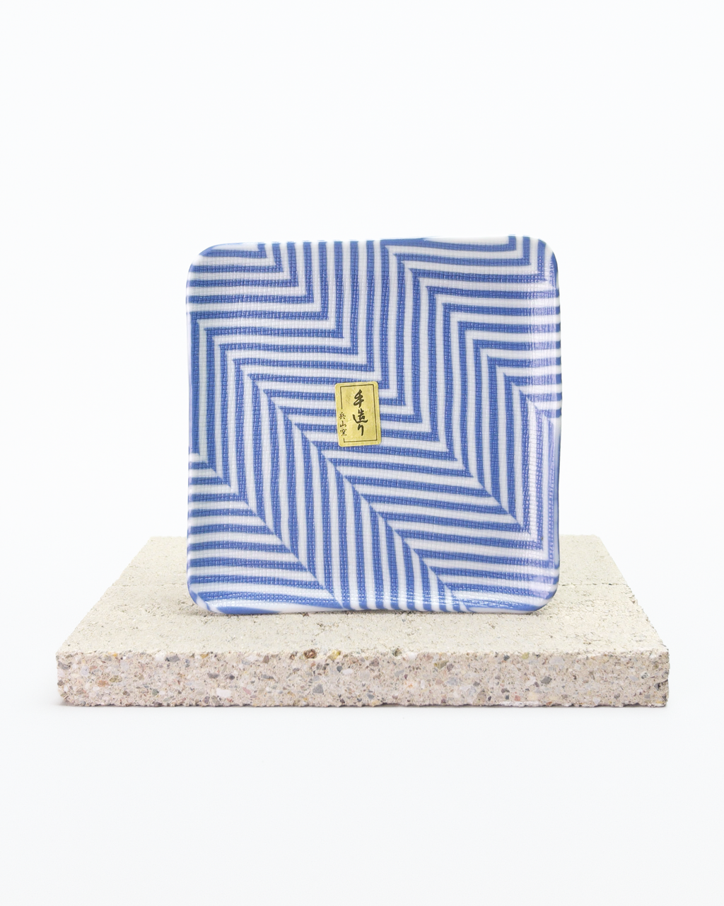 Front side of a blue and white striped nerikomi plate with a gold tag on a beige stone surface