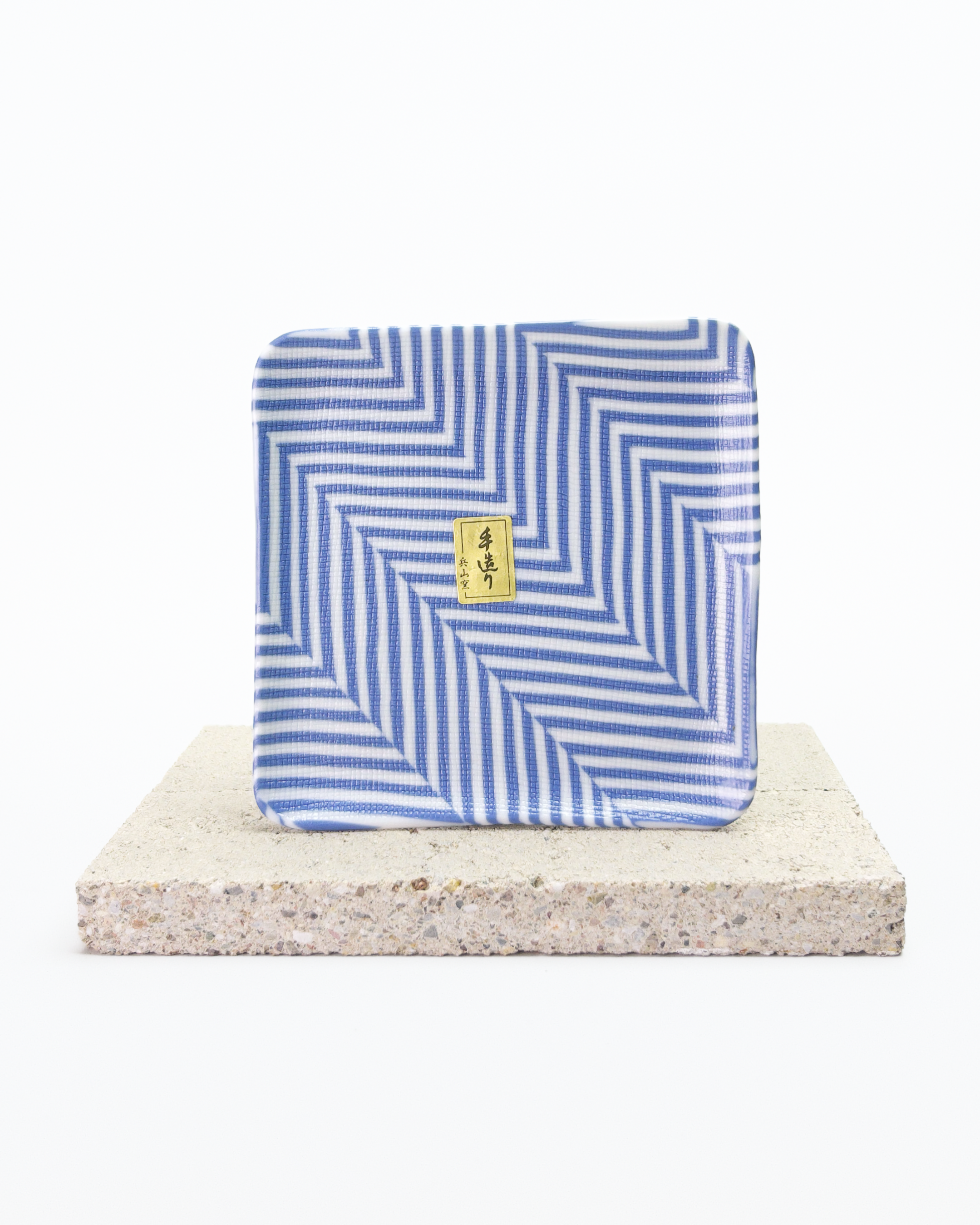 Front side of a blue and white striped nerikomi plate with a gold tag on a beige stone surface